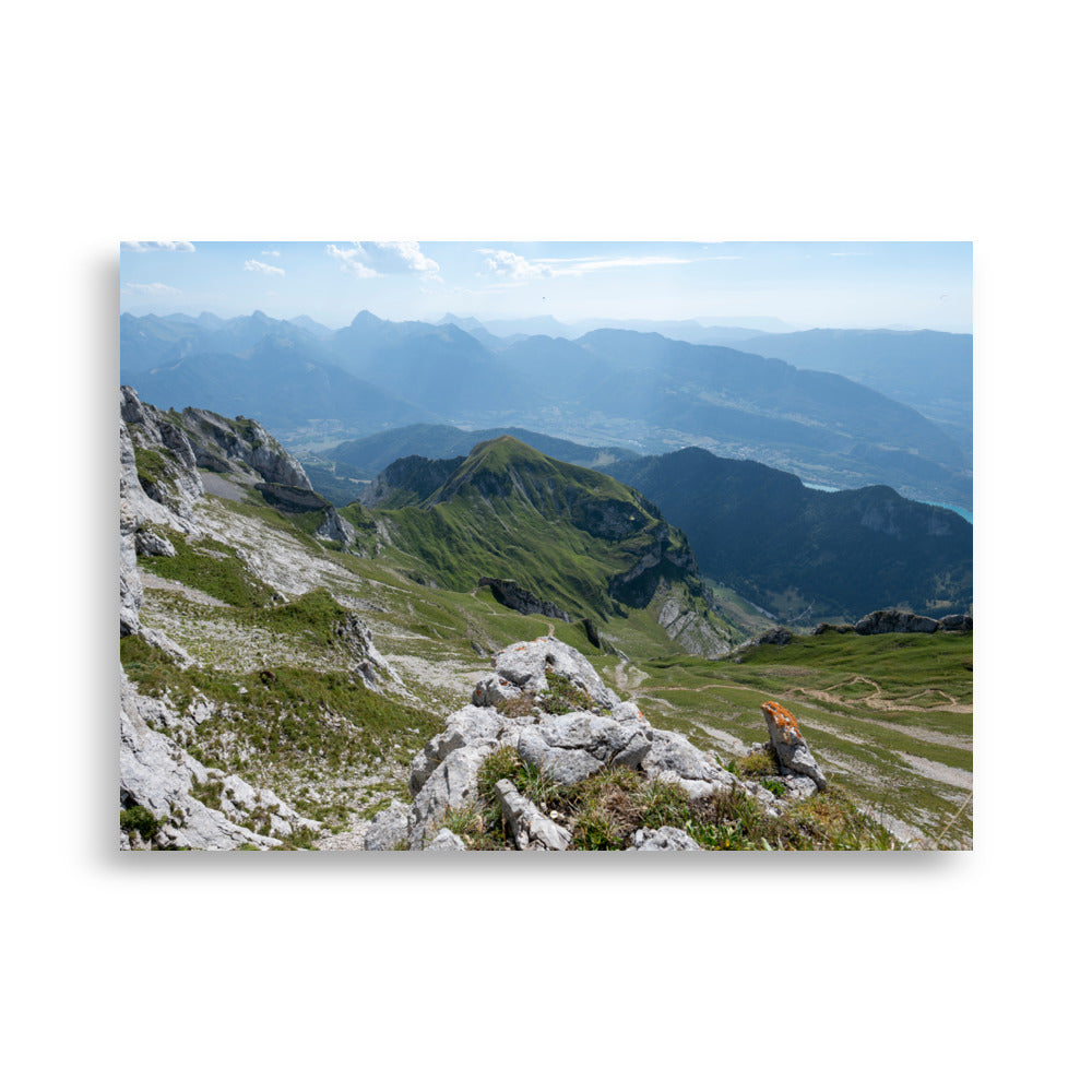 Vue panoramique du lac Annecy depuis les hauteurs, entouré de montagnes majestueuses avec un ciel serein au-dessus. Une représentation fidèle de la beauté naturelle des Alpes.